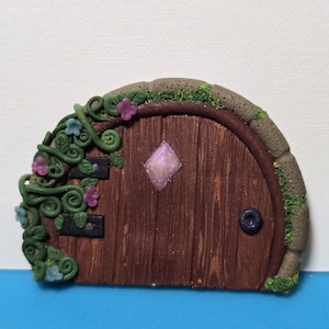 May include: A miniature fairy door with a wooden appearance, featuring a rounded top and a small, sparkling diamond-shaped window. The door is adorned with green vines, small flowers, and a stone-like border, creating a whimsical, fantasy-themed aesthetic.