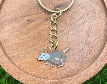 May include: A gold-colored keychain with a cartoon rat wearing a cowboy hat and holding a gun. The keychain is on a wooden coaster.