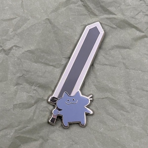 Könnte beinhalten: Eine silberne Emaille-Anstecknadel mit einer Cartoon-Katze, die ein Schwert hält. Die Katze ist hellblau und das Schwert ist schwarz.