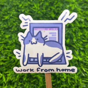 May include: A sticker featuring a cartoon cat sitting in front of a laptop. The cat is predominantly white with blue patches. The laptop screen displays a website interface.  The sticker is purple and white, and the text 'Work From Home' is printed below the illustration. The sticker is mounted on a wooden stick.