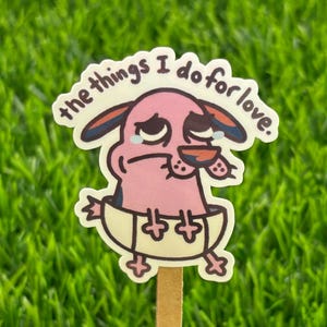 May include: A sticker featuring Courage the Cowardly Dog with the text "the things I do for love." The cartoon dog is pink with blue ears and is sitting in a yellow bowl. The sticker is on a wooden stick, set against a green grass background.