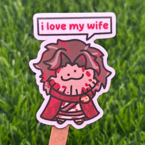 May include: A die-cut sticker featuring a cartoon character with messy brown hair, wearing a red outfit with dark red splatters, and a speech bubble above the head containing the black text "i love my wife" on a white background.