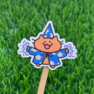 May include: A whimsical sticker featuring an orange cat wearing a blue wizard's hat and robe with yellow stars. The cat has a smiling face and is surrounded by purple swirls. The sticker is attached to a wooden stick, set against a backdrop of green grass.