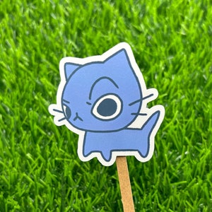May include: A blue cartoon cat sticker with a grumpy expression. The sticker is on a wooden stick and is placed on a green grassy surface.