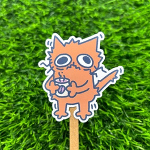 May include: A die-cut sticker featuring a cartoon orange cat with wide eyes and a jagged outline, holding a small white mug with a purple coffee bean graphic. The cat has a distressed expression and is attached to a thin, light brown wooden stick.