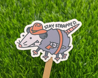 May include: A cartoon sticker of a possum wearing a cowboy hat and holding a gun. The sticker says "Stay Strapped."
