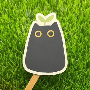 May include: Black cat sticker with a green plant growing out of its head. The cat has yellow eyes and a simple design.