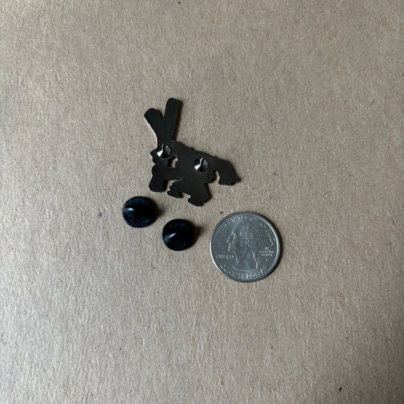 May include: A silver-colored metal pin in the shape of a rabbit, with two black pin backs and a US quarter for size comparison. The rabbit pin has a simple, stylized design.
