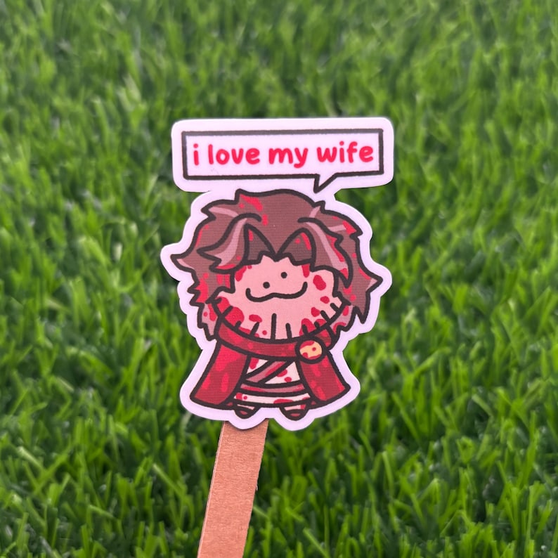 May include: A die-cut sticker featuring a cartoon character with messy brown hair, wearing a red outfit with dark red splatters, and a speech bubble above the head containing the black text "i love my wife" on a white background.
