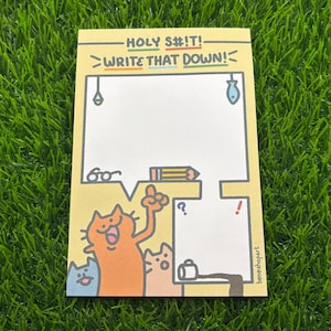Puede incluir: Un bloc de notas amarillo con el texto "HOLY S#!T! WRITE THAT DOWN!" en texto negro y verde. El bloc de notas tiene una ilustración de dibujos animados de tres gatos con un globo de diálogo y un lápiz. El bloc de notas está sobre una superficie verde de hierba.