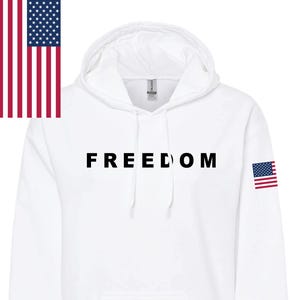 Könnte beinhalten: Weißer Kapuzenpullover mit dem Wort "FREEDOM" in Schwarz auf der Brust. Eine amerikanische Flagge befindet sich auf der linken Seite und am rechten Ärmel. Der Pullover hat eine Vordertasche und eine Kapuze mit Kordelzug.