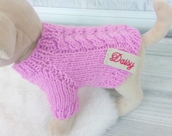 May include: A pink knitted dog sweater with a white tag that says "Daisy" in red embroidery.