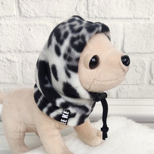 Può includere: Un piccolo cane marrone chiaro che indossa un maglione con cappuccio in pile a stampa leopardo nero e bianco con un cordoncino nero. Il maglione ha la scritta "EEKE" stampata sul lato.