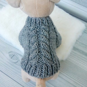 Puede incluir: Un suéter de punto gris para un perro pequeño. El suéter tiene un patrón de punto de cable y está hecho de hilo suave.
