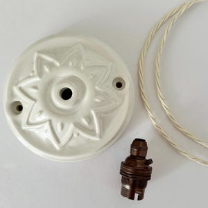 May include: A white ceramic ceiling rose featuring a raised star-shaped floral pattern with two mounting holes, accompanied by a dark metallic light bulb socket and a length of white twisted fabric electrical cord for a pendant light fixture.