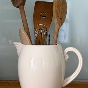 以下が含まれることがあります： 木製のキッチン用品が入った白いセラミックピッチャー。用品には、スプーン、スマイリーフェイスのヘラ、泡立て器が含まれます。ピッチャーは木製の表面に置かれています。