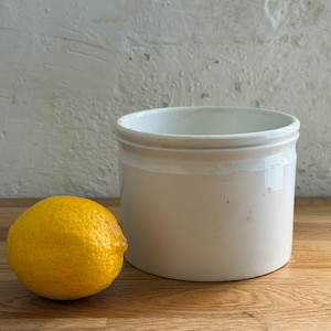 May include: A white ceramic jar with a rounded shape and a wide opening. A bright yellow lemon sits to the left of the jar on a wooden surface. The background is a textured, off-white wall.