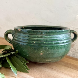 May include: A round, deep green glazed ceramic bowl featuring two small side handles and horizontal ribbed detailing along the exterior. The surface shows subtle variations in color and texture, with small areas of wear along the rim.