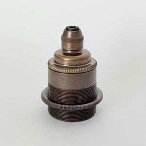 May include: A metallic bronze-toned light bulb socket featuring a cylindrical body with a threaded exterior, a knurled ring detail, and a small black plastic cord grip at the top. The component has a smooth finish and a tiered, industrial design.