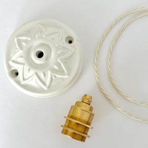 Pode incluir: Uma canopla de teto em cerâmica branca com um design de pétalas florais em relevo, um soquete de lâmpada em latão com anéis roscados e um comprimento de fio elétrico de tecido trançado branco. Os componentes estão dispostos separadamente.