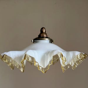 May include: A white glass lamp shade with a ruffled edge and a translucent yellow trim. The shade features a central brass-coloured metal fitter at the top, designed for attachment to a light fixture, showcasing a flared, scalloped silhouette.