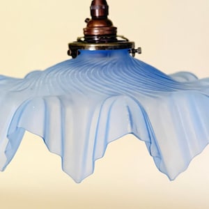 May include: A light blue frosted glass pendant lamp shade with a ruffled, wavy edge and a swirling ribbed pattern. The shade is attached to a metallic bronze-coloured hardware fixture at the top, featuring a central socket and small side screws.