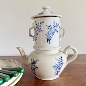 Op de afbeelding: Een witte keramische koffiefilterpot met een blauw bloemmotief. De pot bestaat uit een basis met een tuit en handvat, een middelste filtersectie met twee zijhandvatten en een bijpassend deksel. Een stapel borden met groene handvatten is zichtbaar.