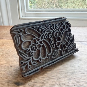 Op de afbeelding: Een rechthoekig houten blok met een uitgesneden bloemenpatroon. Het ontwerp omvat bloemen, bladeren en andere botanische motieven. Het blok heeft een donkere, verouderde uitstraling, met versleten randen. Waarschijnlijk een antiek drukgereedschap.