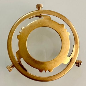 Puede incluir: Un anillo de pantalla de lámpara de metal dorado con tres tornillos. El anillo tiene un diseño decorativo alrededor del borde exterior.