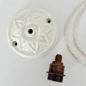May include: A white ceramic ceiling rose featuring a raised floral star pattern, accompanied by a dark bronze metal light bulb socket and a length of white twisted fabric electrical cable for a pendant lighting fixture installation.