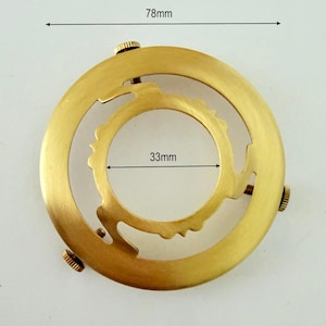 May include: A circular gold-toned metal lamp shade holder featuring a brushed finish. The outer diameter measures 78mm and the inner ring diameter measures 33mm. Three small knurled adjustment screws are spaced evenly around the outer frame.