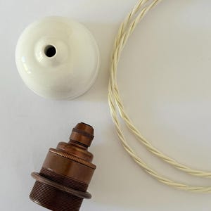 Pode incluir: Uma canopla de teto em cerâmica branca, um soquete de lâmpada em bronze metálico com exterior roscado e um pedaço de fio elétrico de tecido trançado branco. Os componentes estão dispostos separadamente para mostrar o kit de luminária.