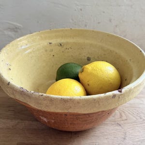 Può includere: Una ciotola in ceramica rotonda con interno e bordo smaltati di giallo, caratterizzata da una base in terracotta naturale. La ciotola contiene due limoni giallo brillante e un lime verde scuro, con una texture maculata sullo smalto.