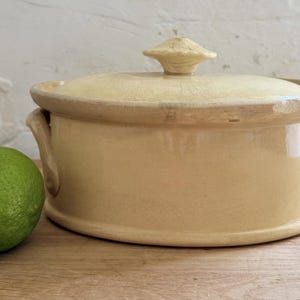 May include: A round, cream-coloured ceramic casserole dish with a matching lid and two side handles. The glaze features a subtle crackle pattern throughout. A single bright green lime is positioned next to the dish for scale reference.
