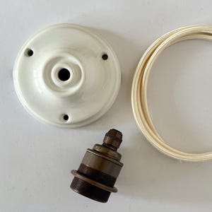 Könnte beinhalten: Ein weißer Deckenbaldachin aus Keramik mit vier Löchern, eine bronzefarbene Lampenfassung und ein cremefarbenes, stoffummanteltes Elektrokabel. Komponenten für eine Leuchte.