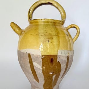 Peut inclure: Une cruche en céramique vernissée avec un corps arrondi, un bec étroit et une grande anse arquée sur le dessus. La partie supérieure est d'un jaune moutarde brillant, tandis que la section inférieure présente une base crème avec des coulures brunes.