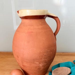 May include: A terracotta pitcher with a rounded body and a single handle. The upper rim and handle feature a cream-coloured glaze with a dark brown band separating it from the natural reddish-brown clay body of the vessel.