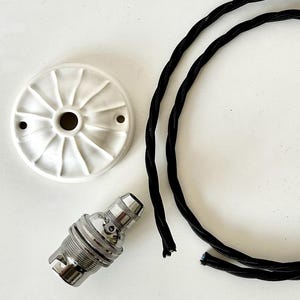 Op de afbeelding: Een bovenaanzicht van een witte keramische plafondkap, een zilverkleurige lamphouder en een zwart gedraaid elektrisch snoer. De kap heeft een cirkelvormig ontwerp met radiale groeven en twee kleine gaten. De lamphouder is cilindrisch met een geribbelde textuur.