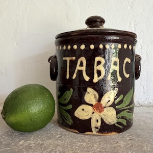 Può includere: Un barattolo in ceramica marrone scuro con coperchio e due manici laterali, caratterizzato dalla scritta TABAC in lettere color crema e da un motivo floreale dipinto a mano con foglie verdi e un fiore color crema. Un lime verde è accanto.