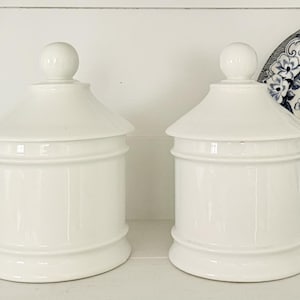 Può includere: Due barattoli in ceramica bianca identici con corpi cilindrici, basi svasate e coperchi abbinati sormontati da pomelli sferici. I barattoli presentano una finitura liscia e lucida e un design pulito e semplice.