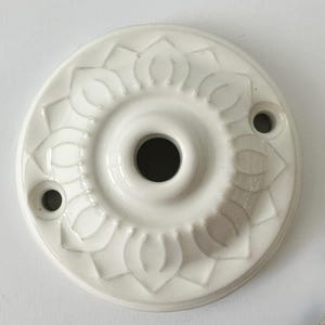Può includere: Una rosetta da soffitto in ceramica bianca con un motivo floreale. La rosetta rotonda ha un foro centrale e due fori più piccoli per il montaggio. La superficie è lucida.