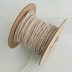 Puede incluir: Una bobina de cordón trenzado beige. El cordón está enrollado alrededor de una bobina de cartón con un núcleo de metal plateado. El cordón tiene un aspecto texturizado y es adecuado para manualidades, fabricación de joyas o fines decorativos.