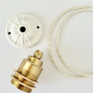 Pode incluir: Uma canopla de teto de cerâmica branca com padrão radial canelado, um soquete de lâmpada de latão com anéis roscados e um pedaço de fio elétrico de tecido trançado branco. Os componentes estão dispostos separadamente para mostrar as peças.