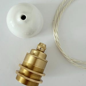 Pode incluir: Um bocal de latão com exterior roscado, uma canopla de teto de cerâmica branca com uma abertura central e um fio elétrico de tecido trançado branco. Os componentes estão dispostos separadamente para mostrar as peças individuais.