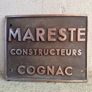 Könnte beinhalten: Eine rechteckige, bronzefarbene Metallplatte mit erhabener Beschriftung. Der Text lautet MARESTE in der oberen Zeile, CONSTRUCTEURS in der mittleren Zeile und COGNAC in der unteren Zeile. Die Platte weist vier kleine kreisförmige Löcher auf.
