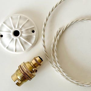 May include: A white ceramic ceiling rose with a brass lamp holder and a silver and white twisted cable. The components are arranged on a white surface. The ceiling rose has a diameter of approximately 10 cm.