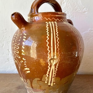 May include: A brown glazed ceramic pitcher with a rounded body, a single arched handle on top, and a small spout. The exterior features vertical cream-coloured slip-trailed patterns consisting of wavy lines and floral motifs around the vessel.