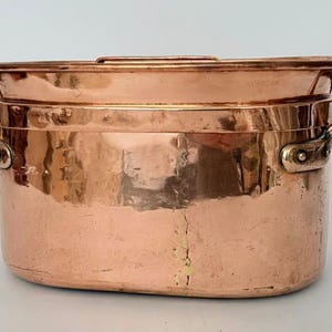 May include: An oval-shaped copper pot with a hammered finish and a matching lid. The pot features two riveted side handles made of the same copper material. The surface shows a warm metallic sheen with visible signs of wear and texture.