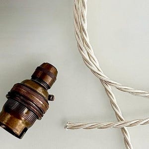Peut inclure: Une douille de lampe en laiton de style vintage avec un câble électrique torsadé argenté. La douille a une forme cylindrique avec un design nervuré. Le câble est composé de plusieurs brins argentés torsadés.