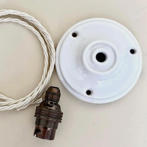 Op de afbeelding: Een witte keramische plafondkap met drie montagegaten, een bronzen lamphouder en een wit en grijs gedraaid elektrisch snoer. Componenten voor een armatuur.